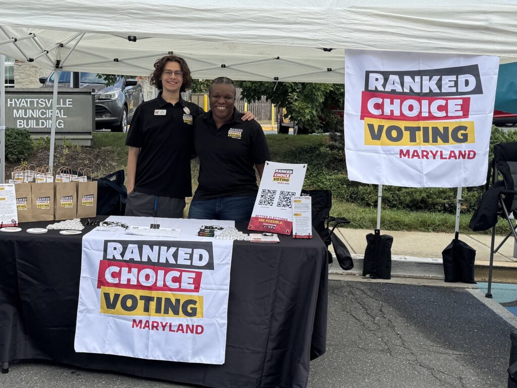 Owen (Deputy Director) and Michelle (Executive Director) smiling while standing at a table with banners reading "Ranked Choice Voting Maryland"